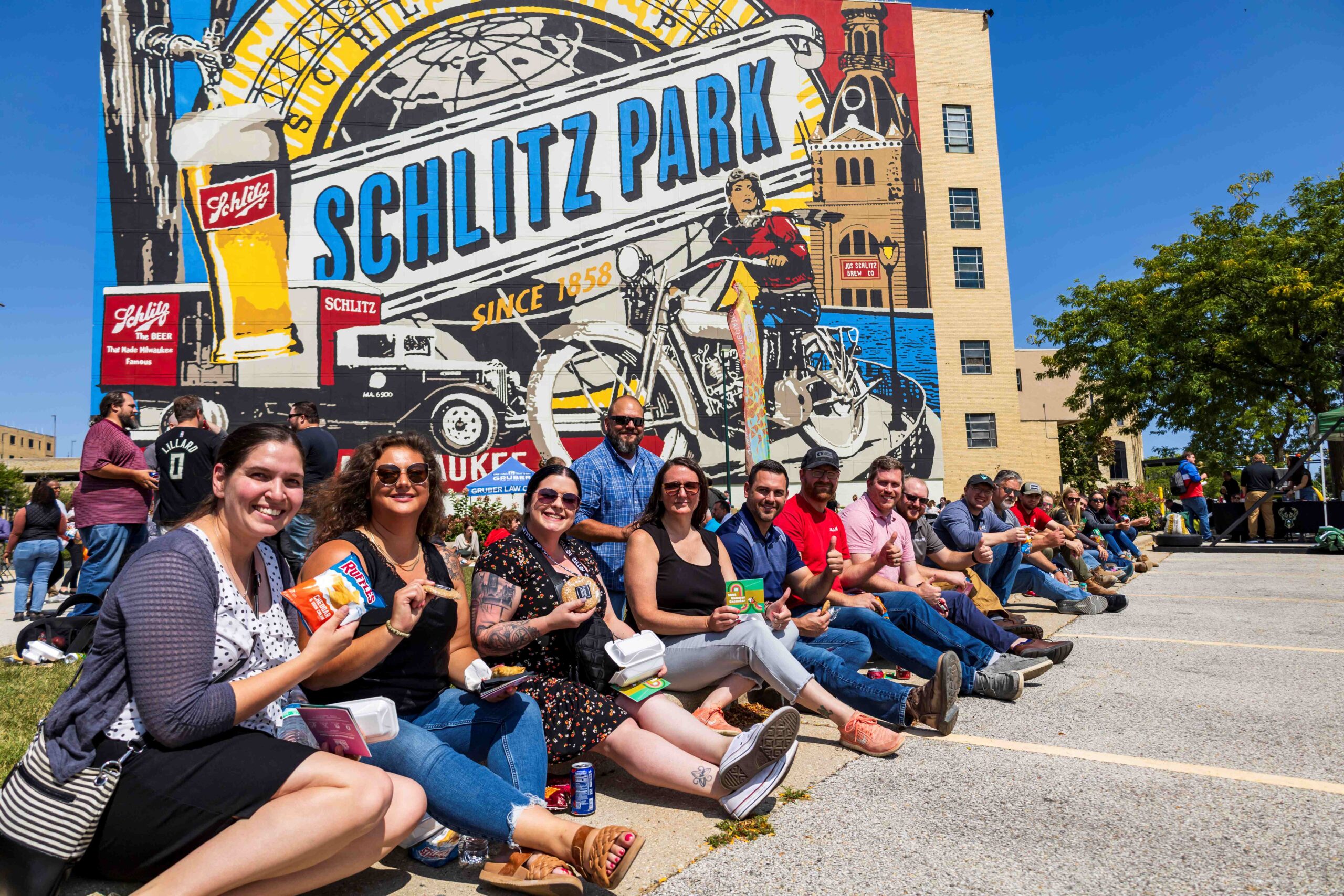 A group of people sits on the curb in from the Schlitz Park Mural.